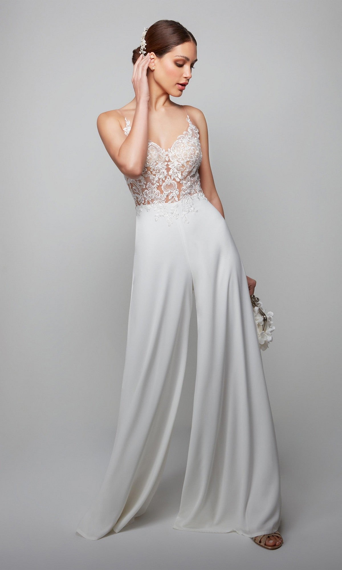 A woman in the Alyce 70017 long ivory jumpsuit, featuring a white lace bodice and wide-leg pants, holds a small bouquet while standing against a plain light gray background.